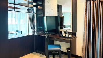 A hotel room with a desk, a television, a chair, a mirror, and a window with curtains.
