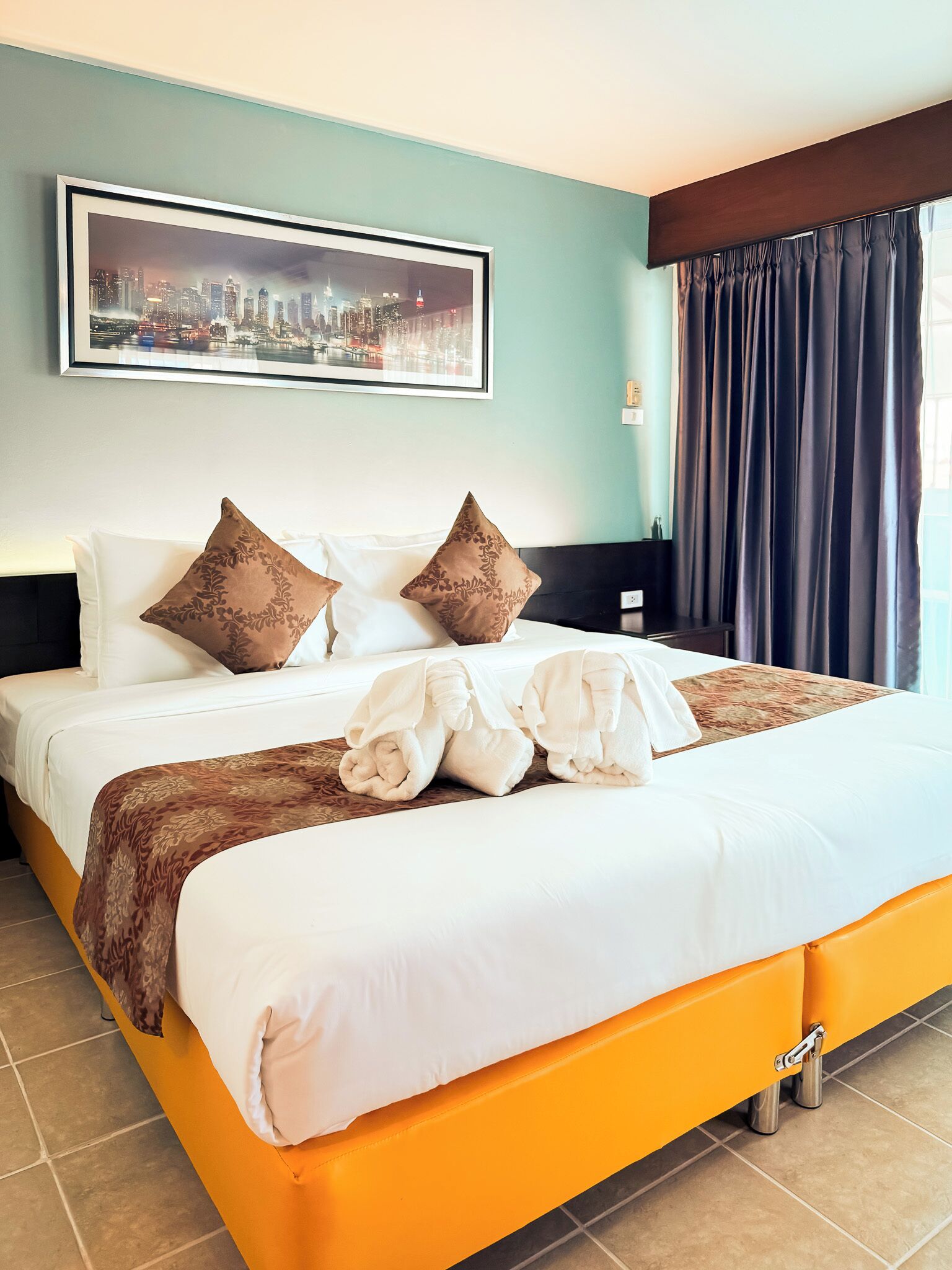 A hotel room with a bed, pillows, a blanket, a cityscape painting, and a window with curtains.