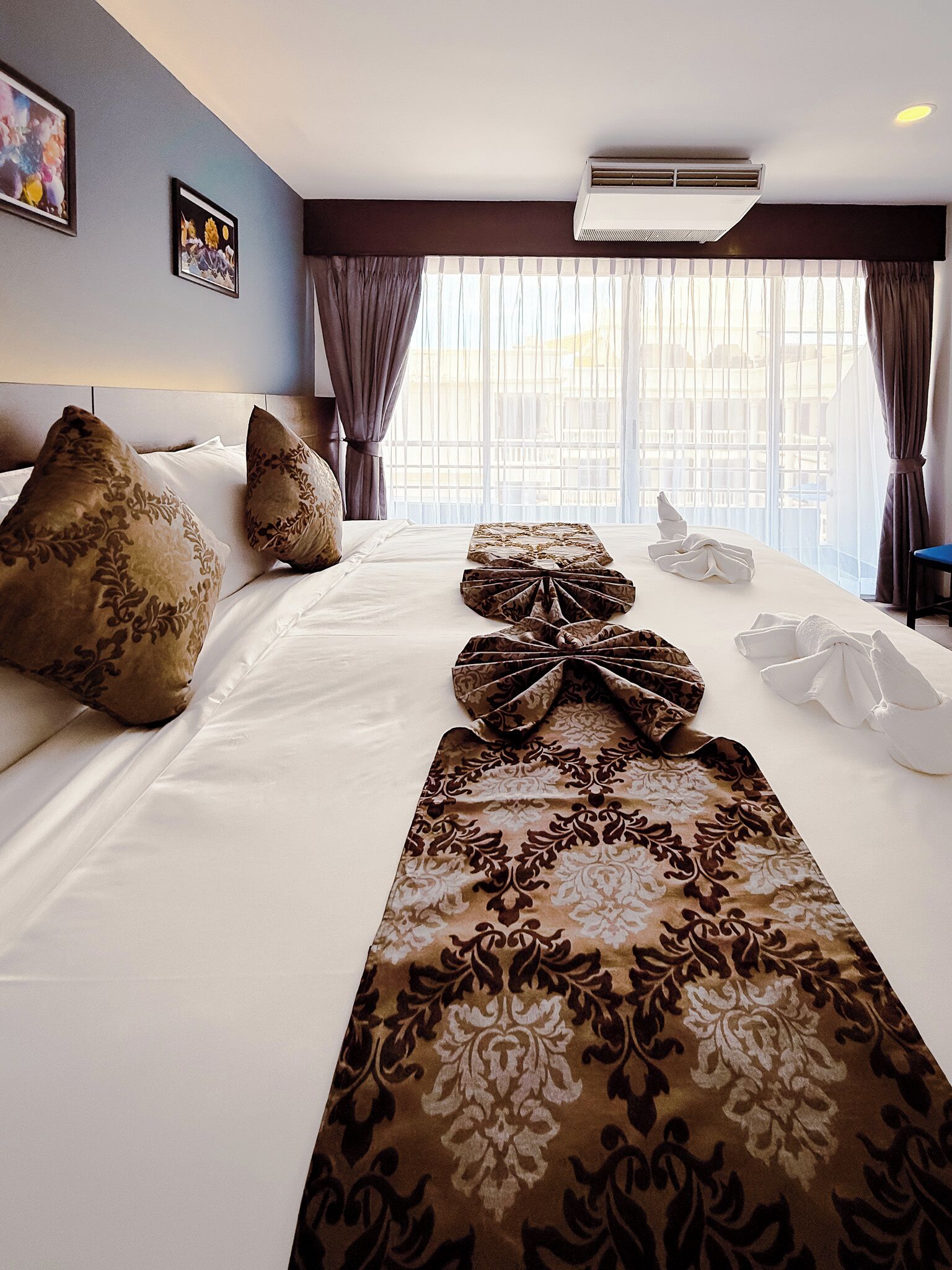 A hotel room with a large bed, a headboard, a window with curtains, and an air conditioning unit.