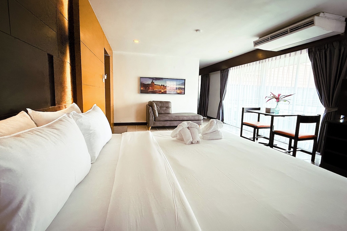 A hotel room with a large bed, a small table with chairs, a painting on the wall, and a window with curtains.