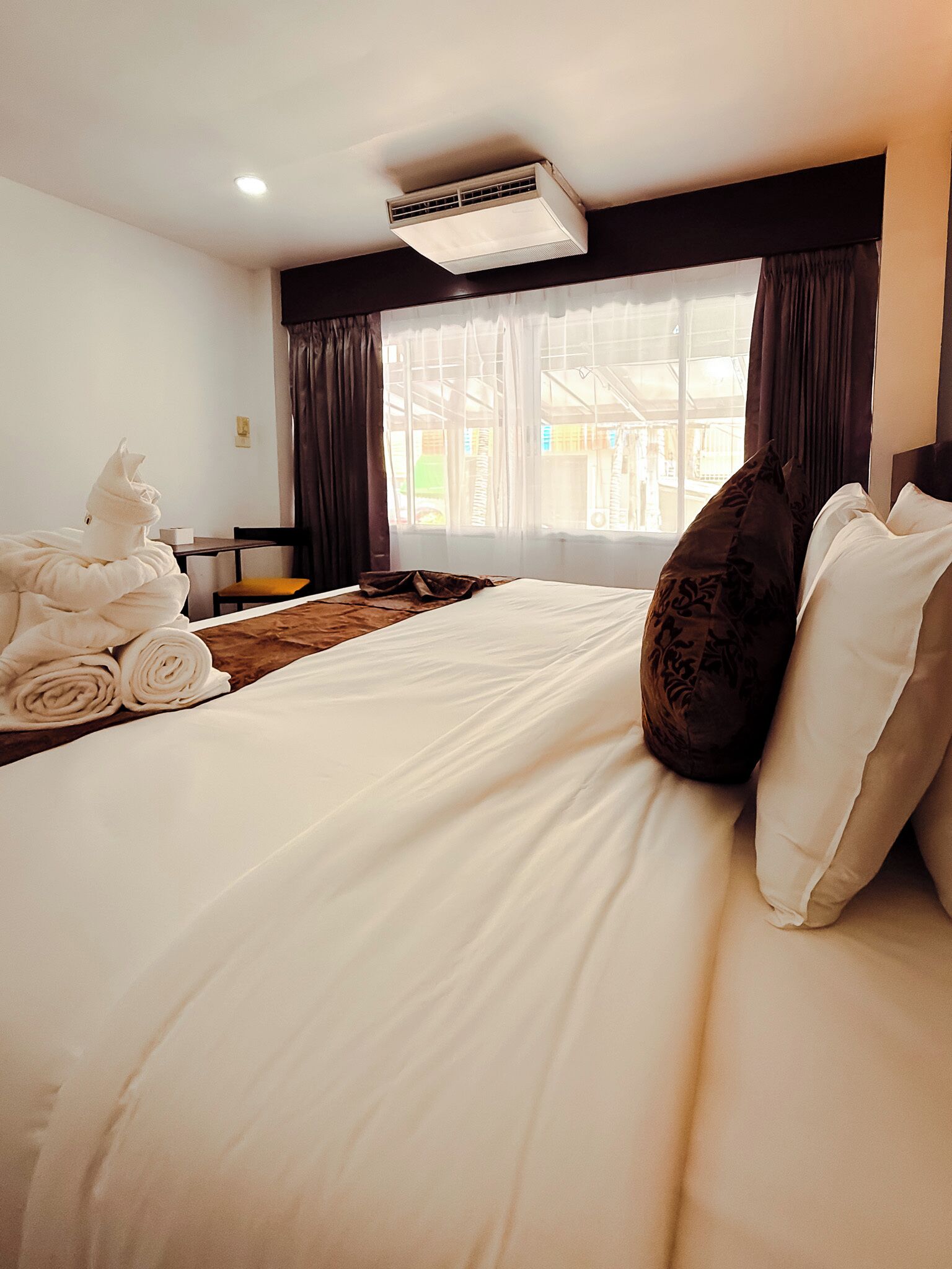 A hotel room with a bed, a window with curtains, an air conditioning unit, and a chair.