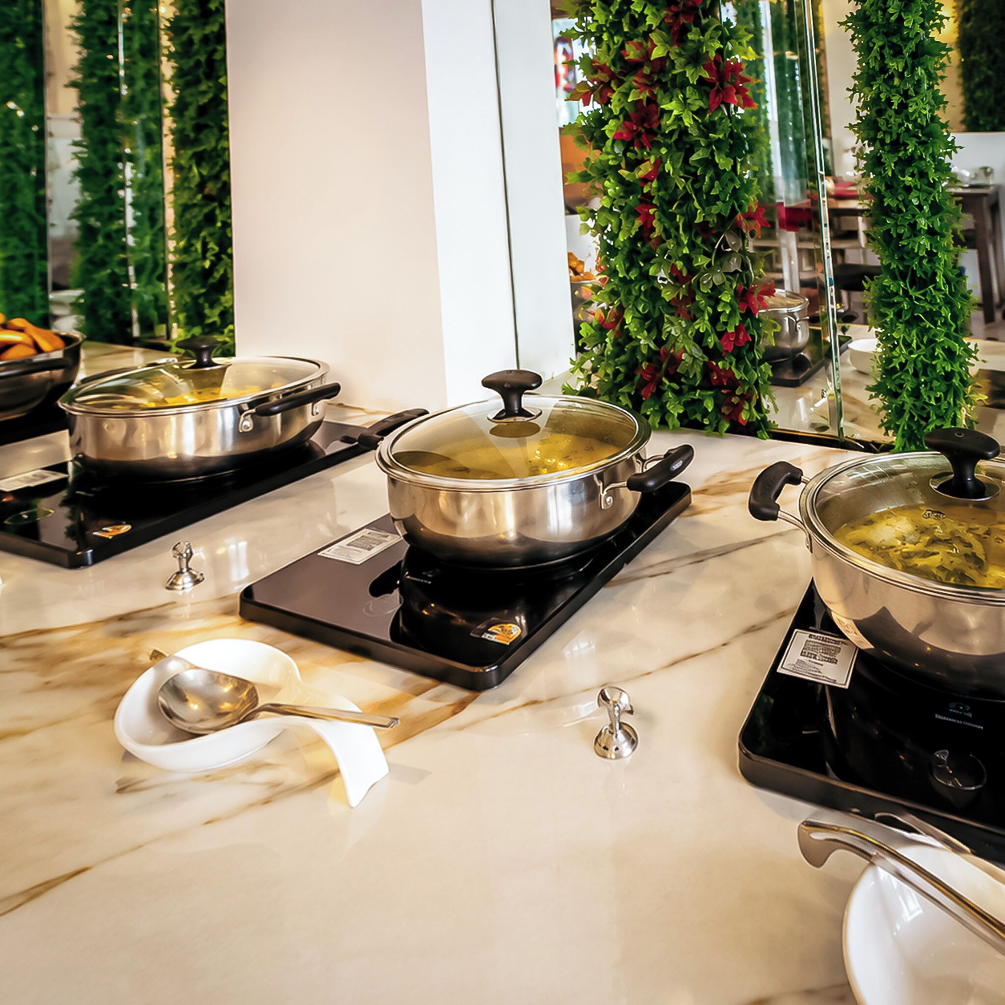 A kitchen area with three pots on electric stoves, a white marble countertop, and decorative greenery in the background.
