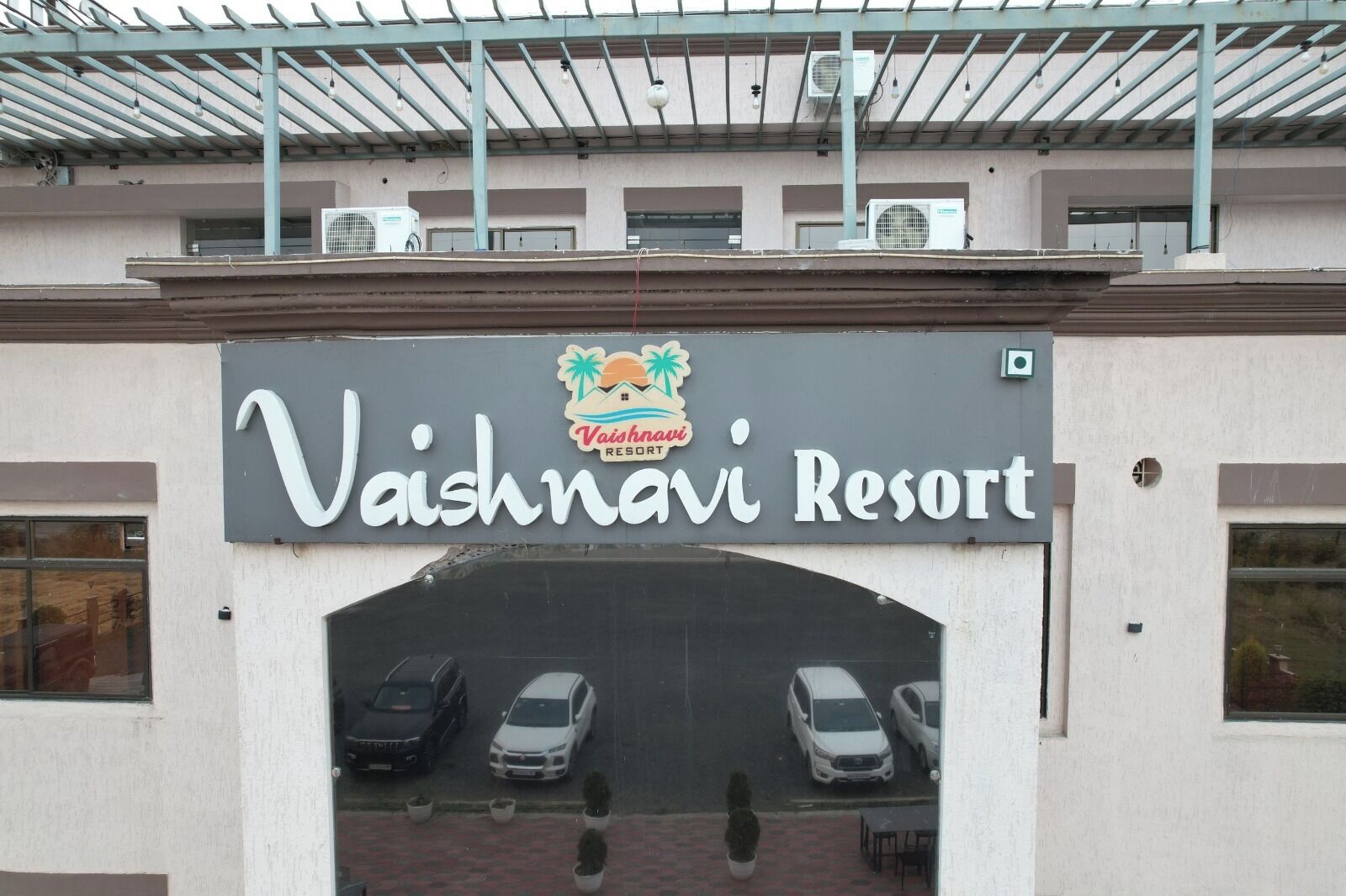 A building with a sign for Vaishnavi Resort, featuring a covered parking area with several cars.
