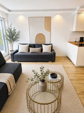 Ein modernes Wohnzimmer mit einer Couch, einem Couchtisch mit einer Pflanze und einer Küchenzeile mit einem Wasserkocher.