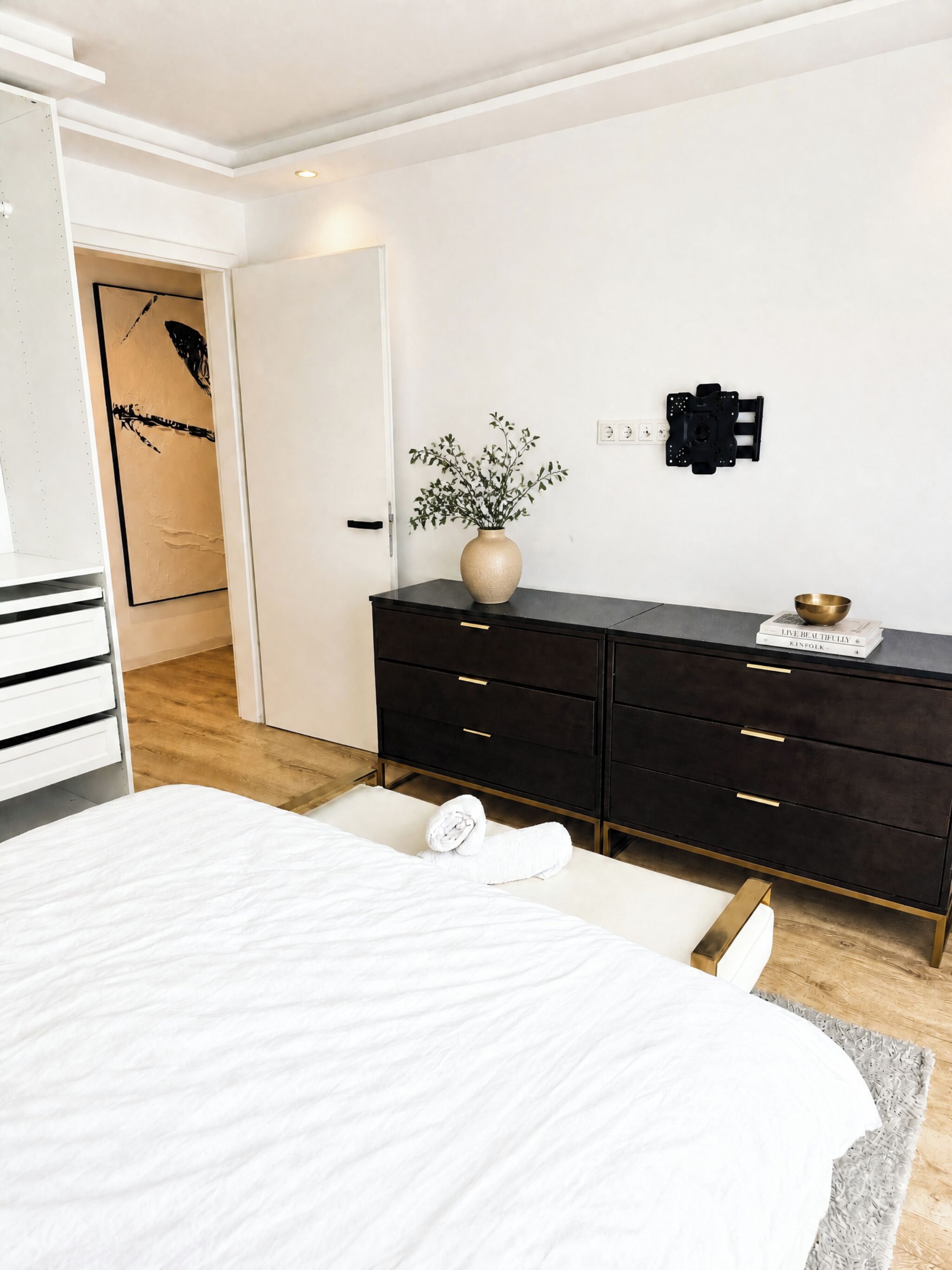 Ein Schlafzimmer mit einem Bett, einer Kommode, einer Vase mit Blumen und einem Wandbild.