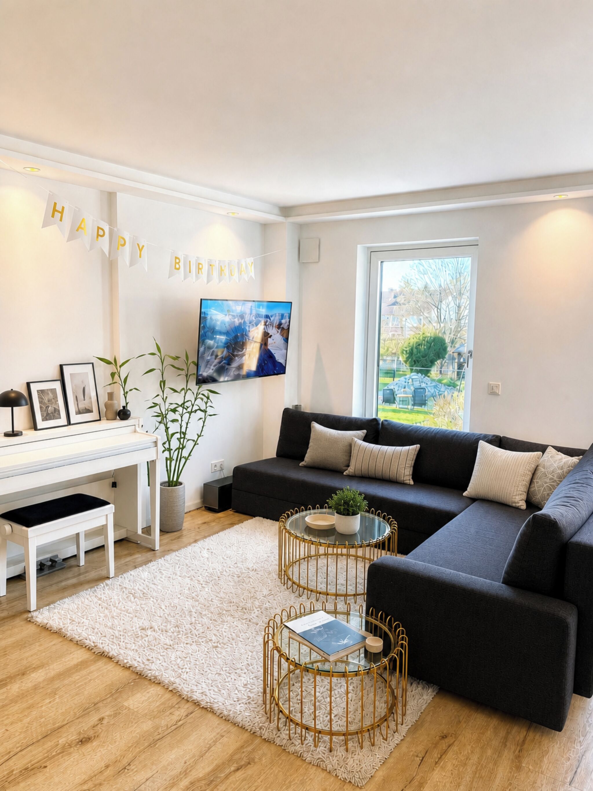 A modern living room with a black sofa, a white piano, a glass coffee table, and a 'HAPPY BIRTHDAY' banner.