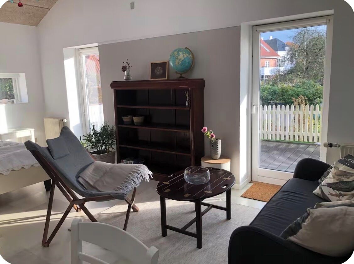 A living room with a sofa, a chair, a coffee table, a bookshelf, and a window with a view of a white fence and greenery.