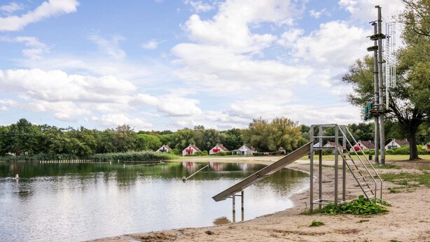 Una zona a la vora d'un llac amb un tobogan metàl·lic, una platja de sorra i cases al fons.