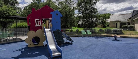 A playground with a slide and climbing structures, surrounded by a fenced area with trees and a building in the background.