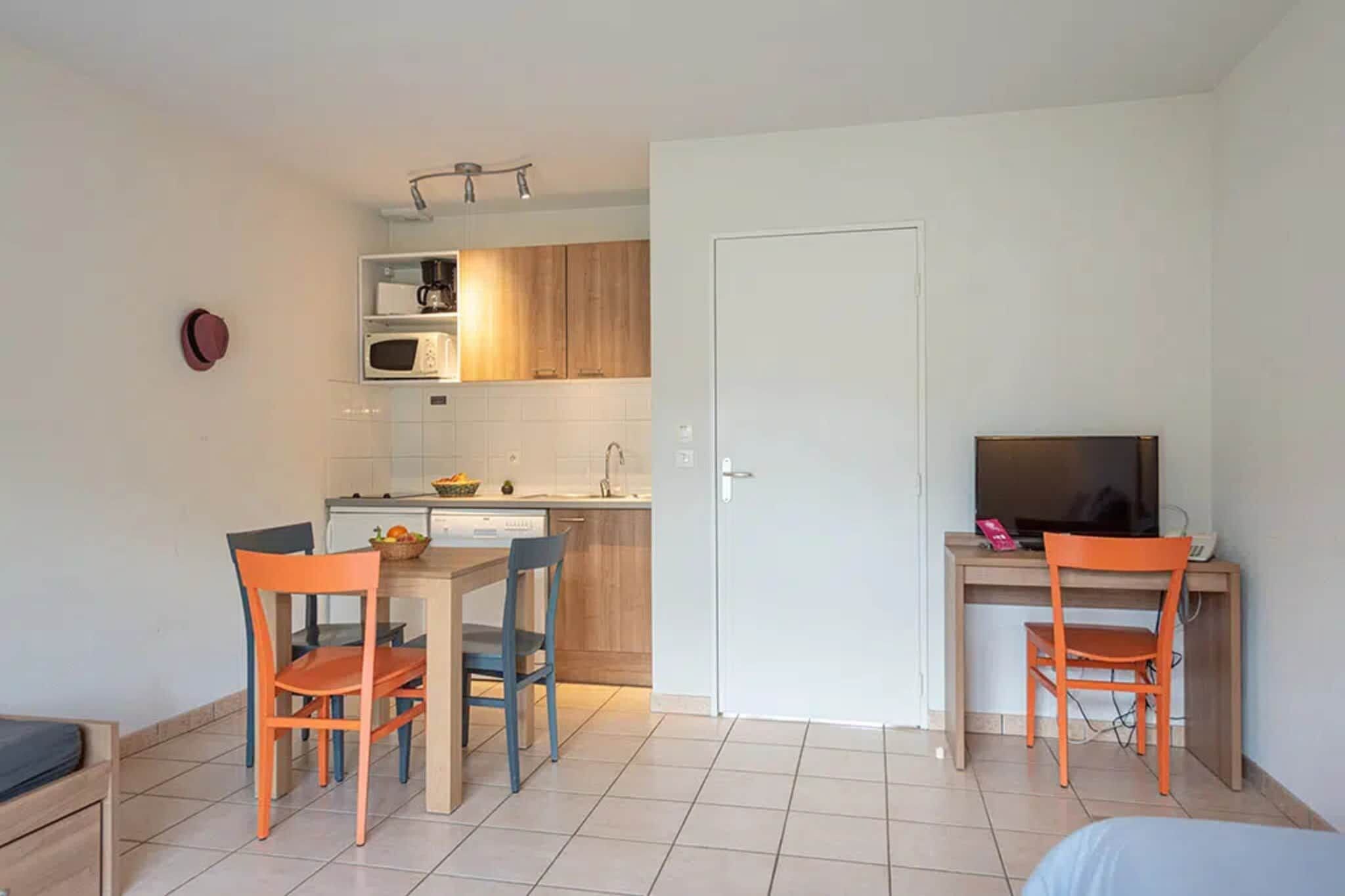 Een kleine keuken met een magnetron, spoelbak en houten kastjes. Er is een eethoek met twee stoelen en een tafel. Een televisie staat op een bureau naast de eethoek.