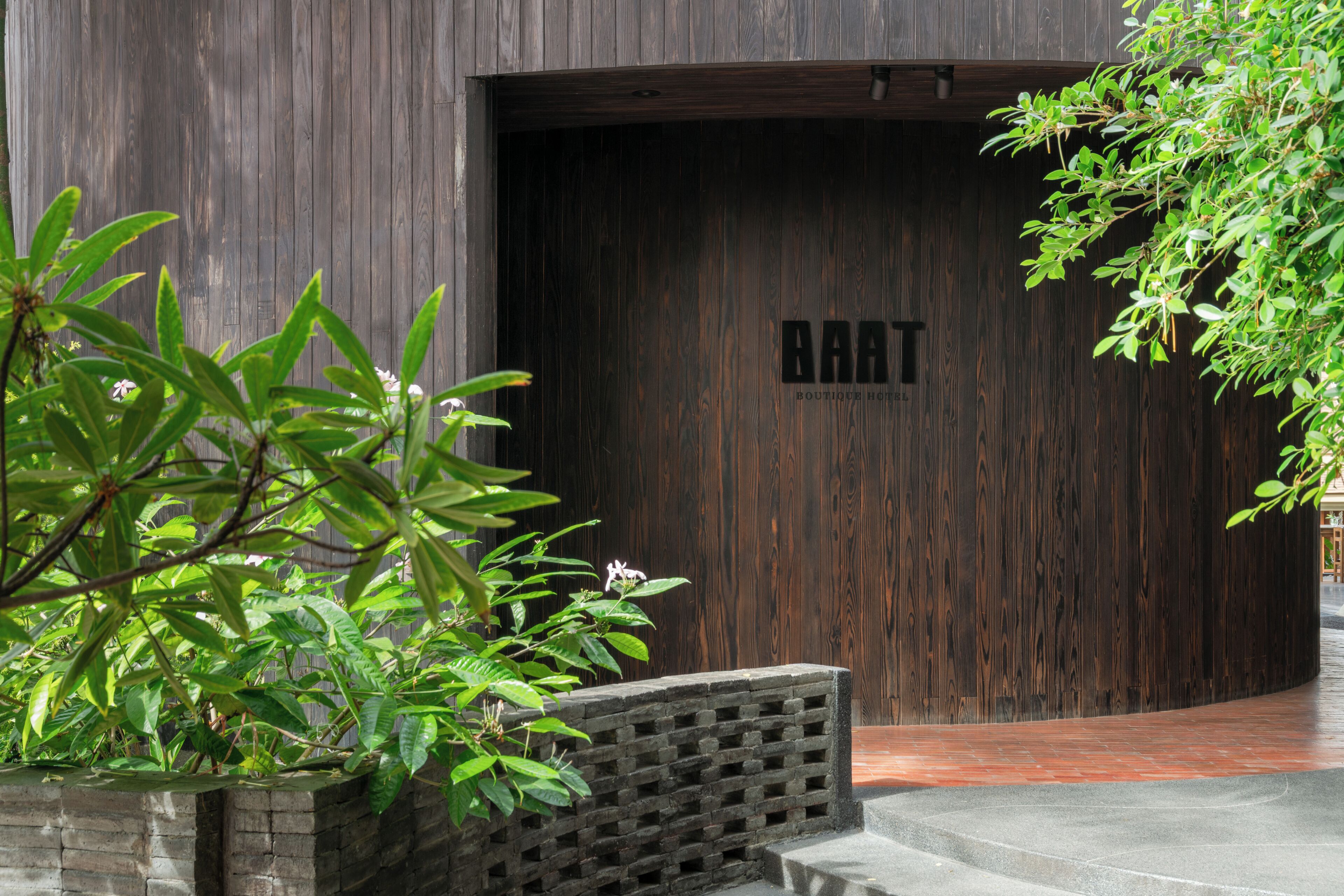Una puerta de madera con el texto 'BAAT' y una jardinera de ladrillo con follaje verde.