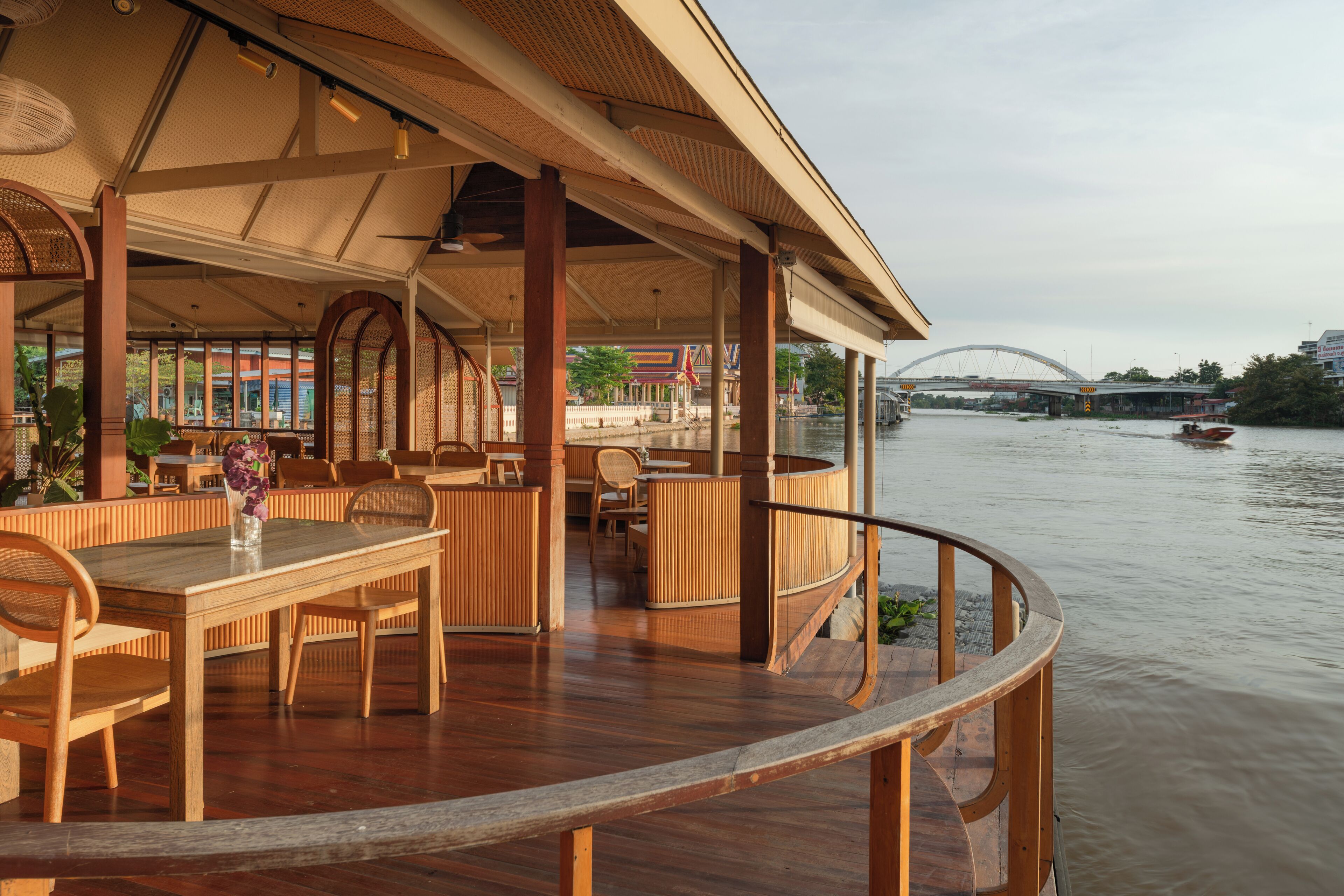 A wooden deck with tables and chairs, overlooking a river with a bridge in the background.
