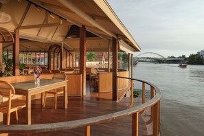 Una terraza de madera con mesas y sillas, con vista a un río y un puente al fondo.