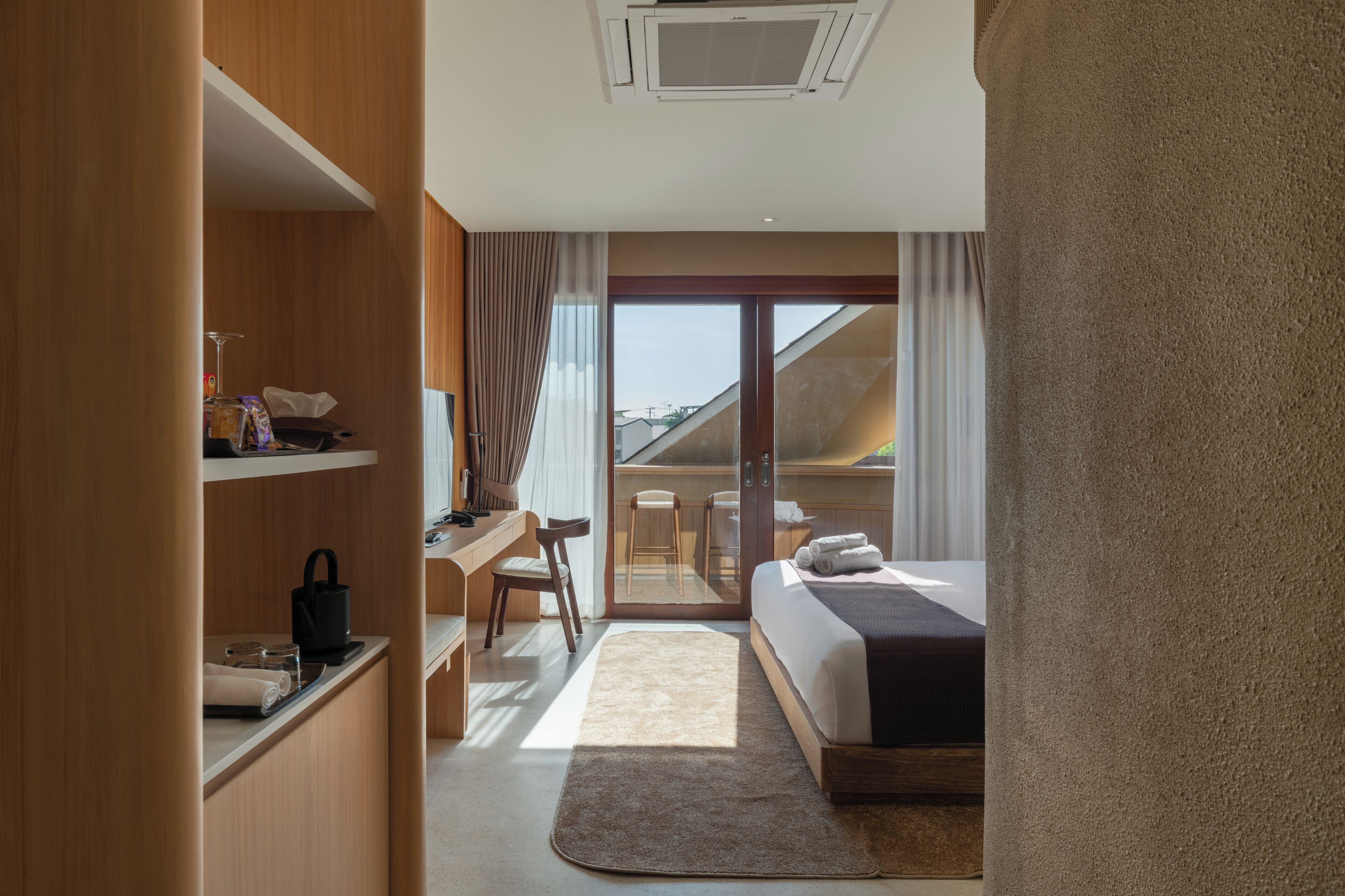 A hotel room with a bed, a desk, a chair, a small kitchenette, and a sliding glass door leading to a balcony.