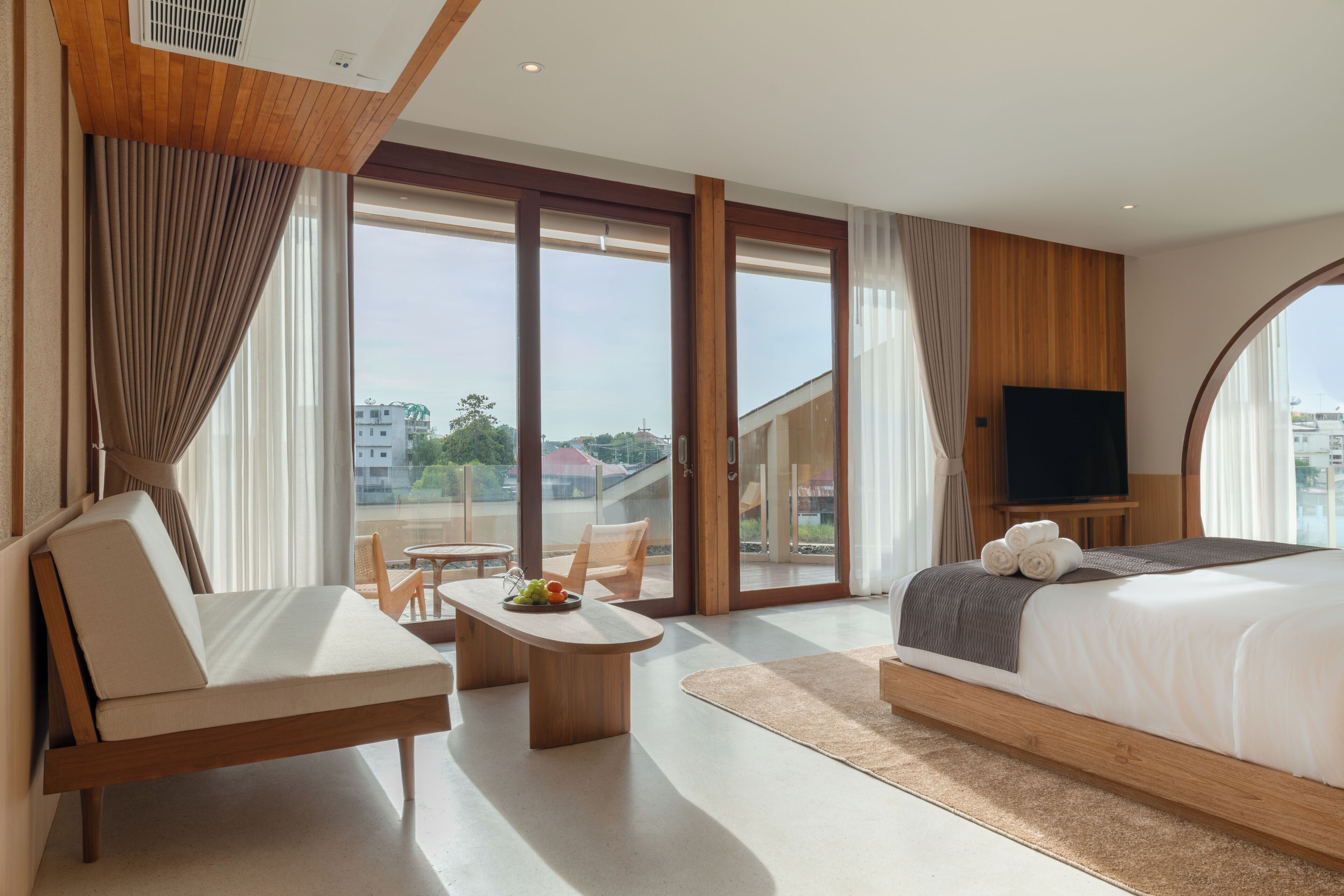 A hotel room with a bed, a TV, a seating area with a table, and large windows offering a view of the city.
