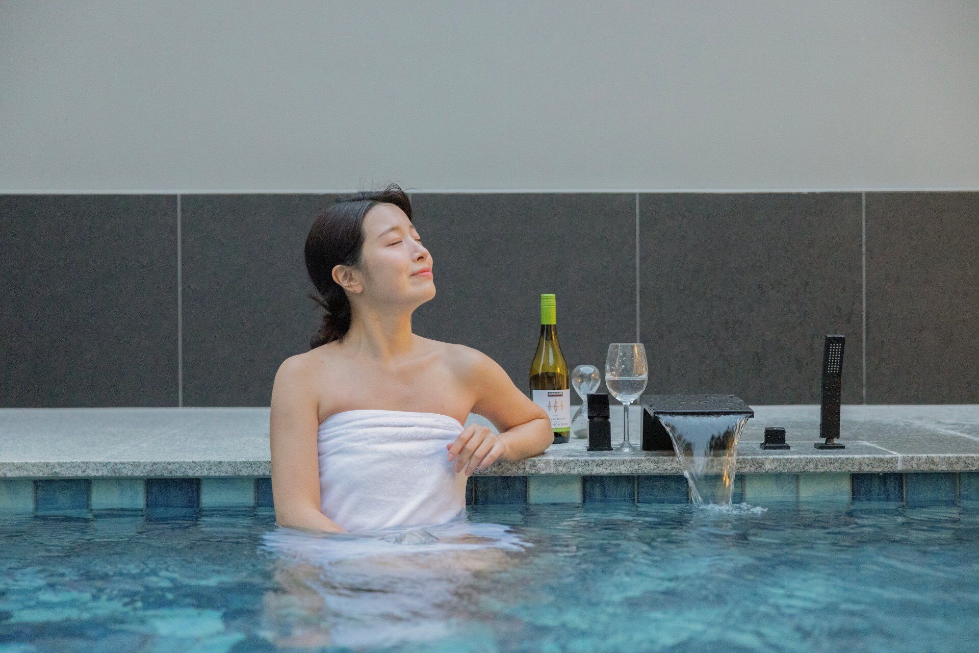 A person in a swimming pool with a towel wrapped around their body, a bottle of wine, and a glass on the poolside.