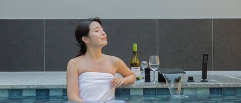 A person in a swimming pool with a towel wrapped around their body, a bottle of wine, and a glass on the poolside.
