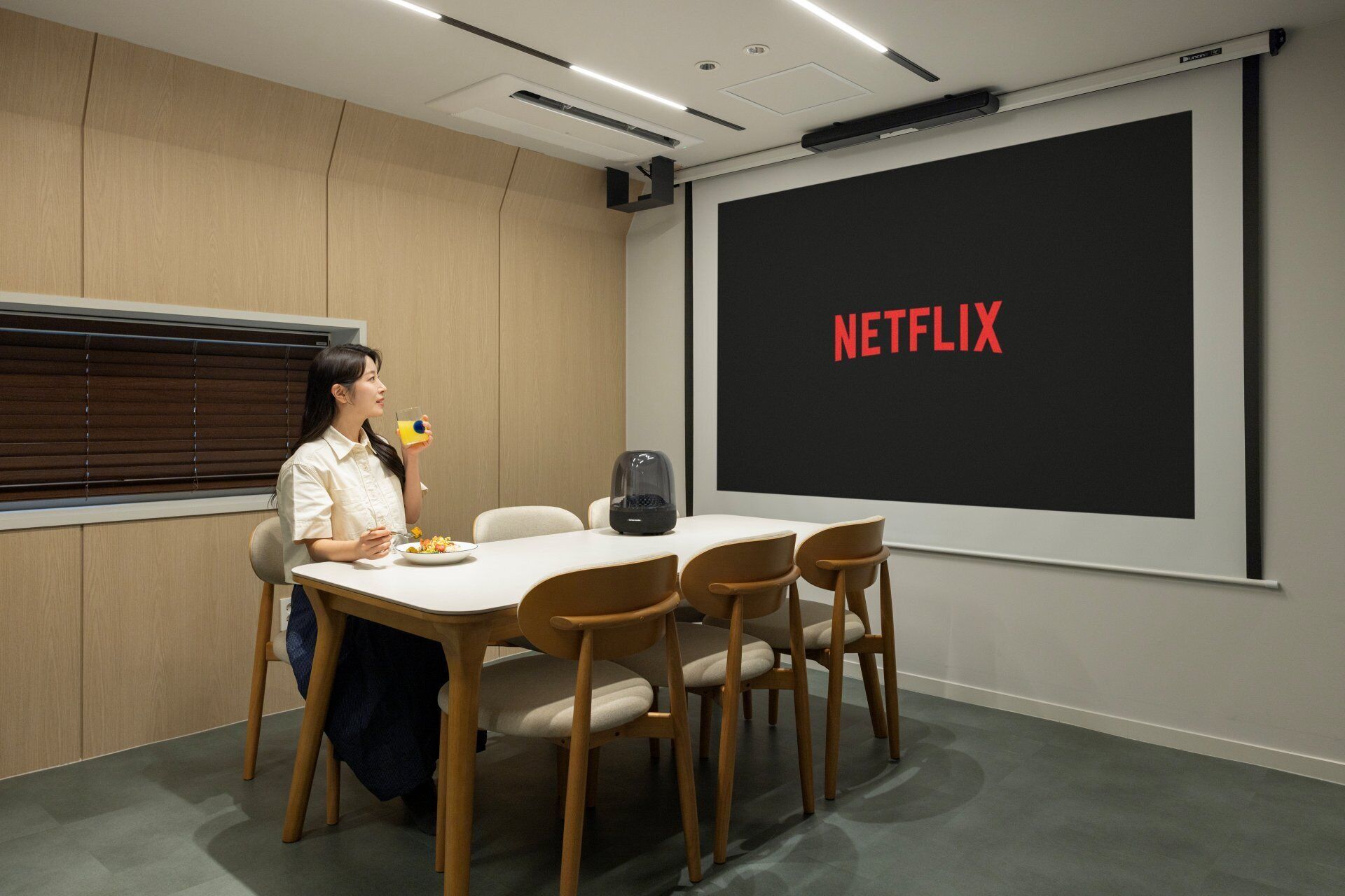 A conference room with a large screen displaying 'NETFLIX,' a table with chairs, and a person holding a drink.