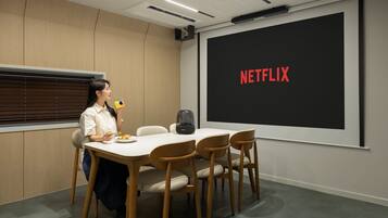 Una sala de conferencias con una pantalla grande que muestra 'NETFLIX', una mesa con sillas y una persona sosteniendo una bebida.