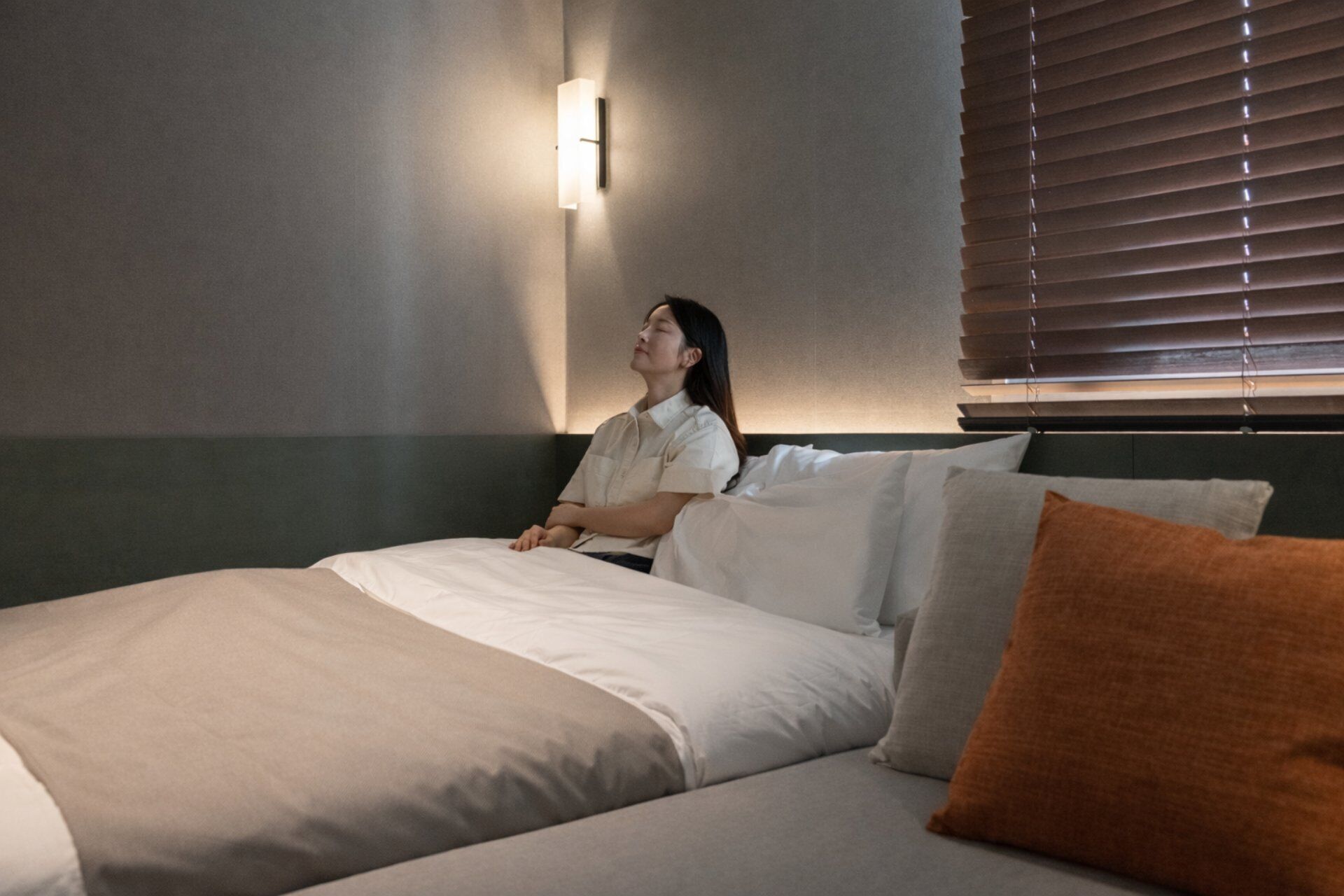 A hotel room with a bed, pillows, a wall-mounted light, and a window with blinds.