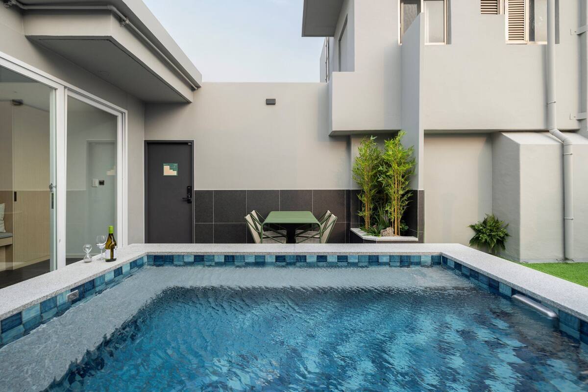 Una piscina con borde de azulejos, una puerta con cerradura, una pequeña mesa con sillas y una planta en maceta.