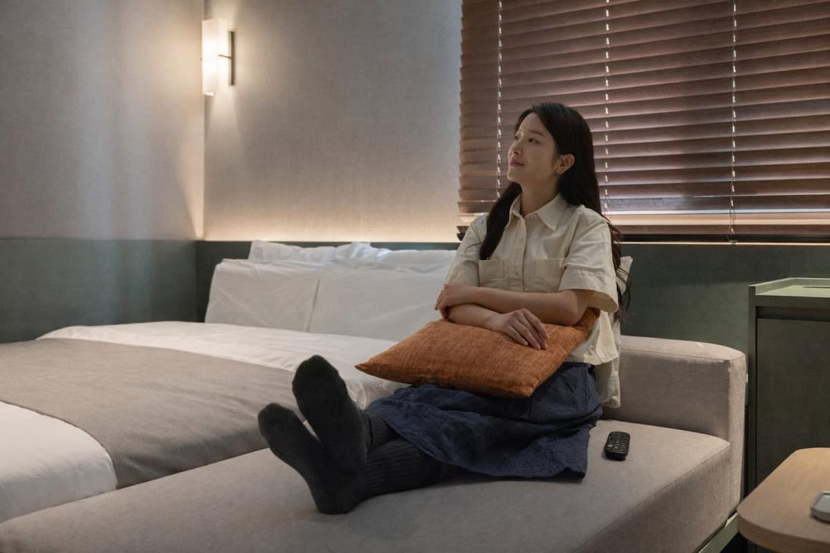 Una persona sentada en una cama con un almohadón, un control remoto sobre la cama y una mesita de noche con una lámpara.
