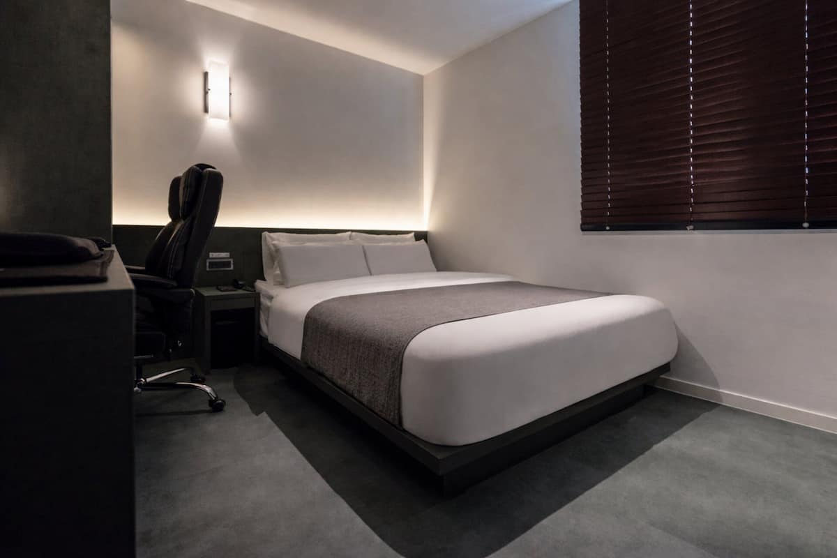 Una habitación de hotel moderna con una cama, un escritorio, una silla y una lámpara fijada a la pared.