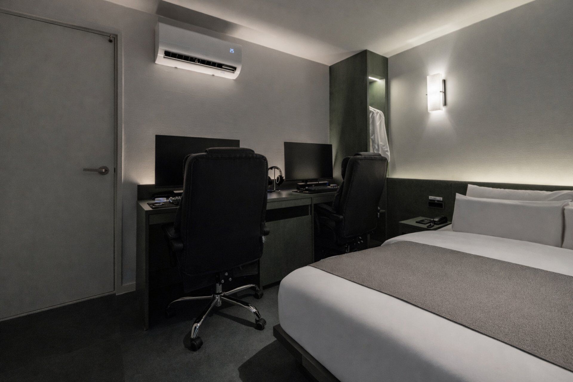 A hotel room with a bed, two desks with computers, an air conditioner, and a nightstand with a lamp.
