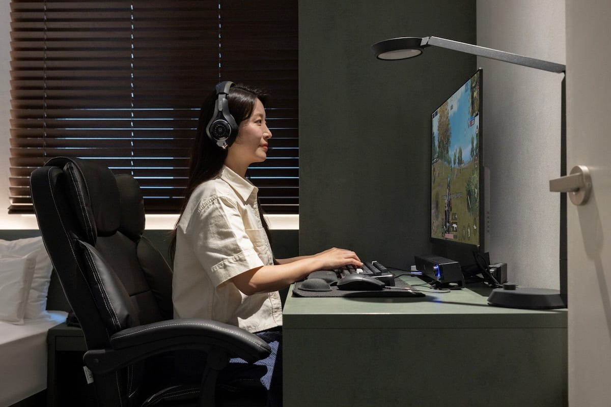 Una persona sentada en un escritorio con una computadora, audífonos y un monitor que muestra un videojuego.