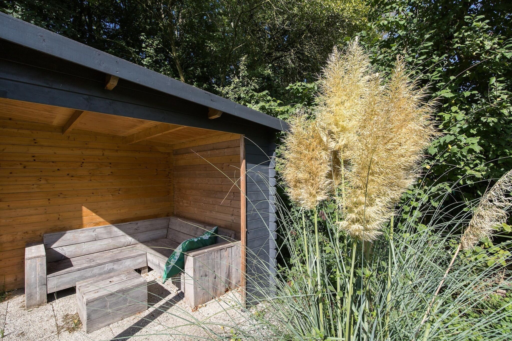 Een houten zitgedeelte buiten met banken en een tafel, omgeven door hoge grassen en bomen.