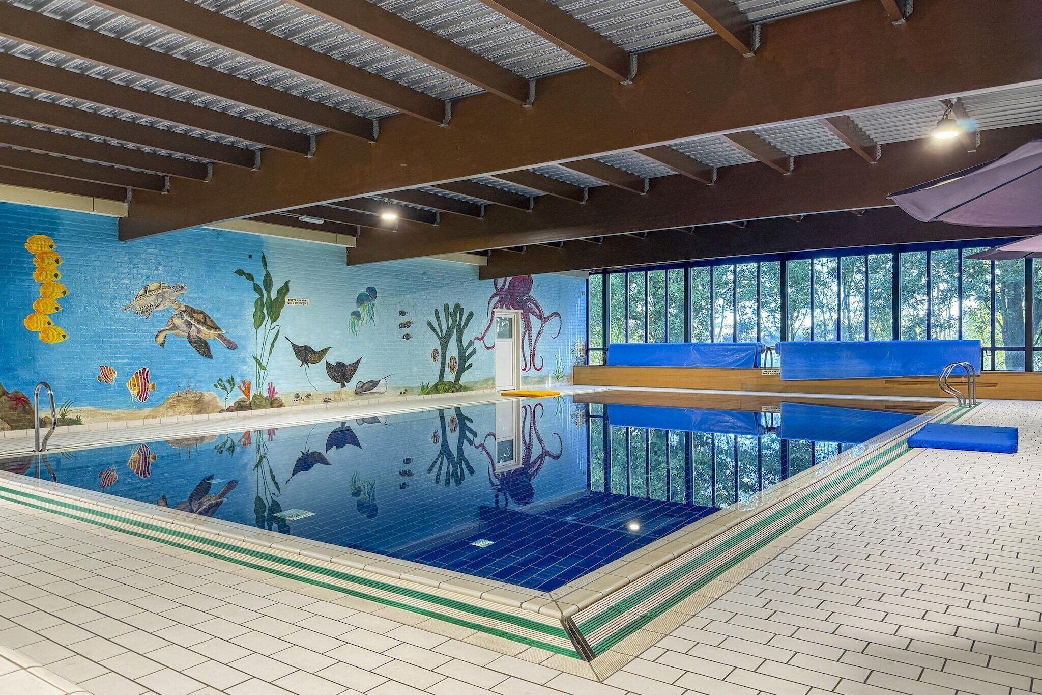 An indoor swimming pool with a mural of aquatic life, a diving board, and a view of trees through large windows.