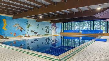An indoor swimming pool with a mural of aquatic life, a diving board, and a view of trees through large windows.