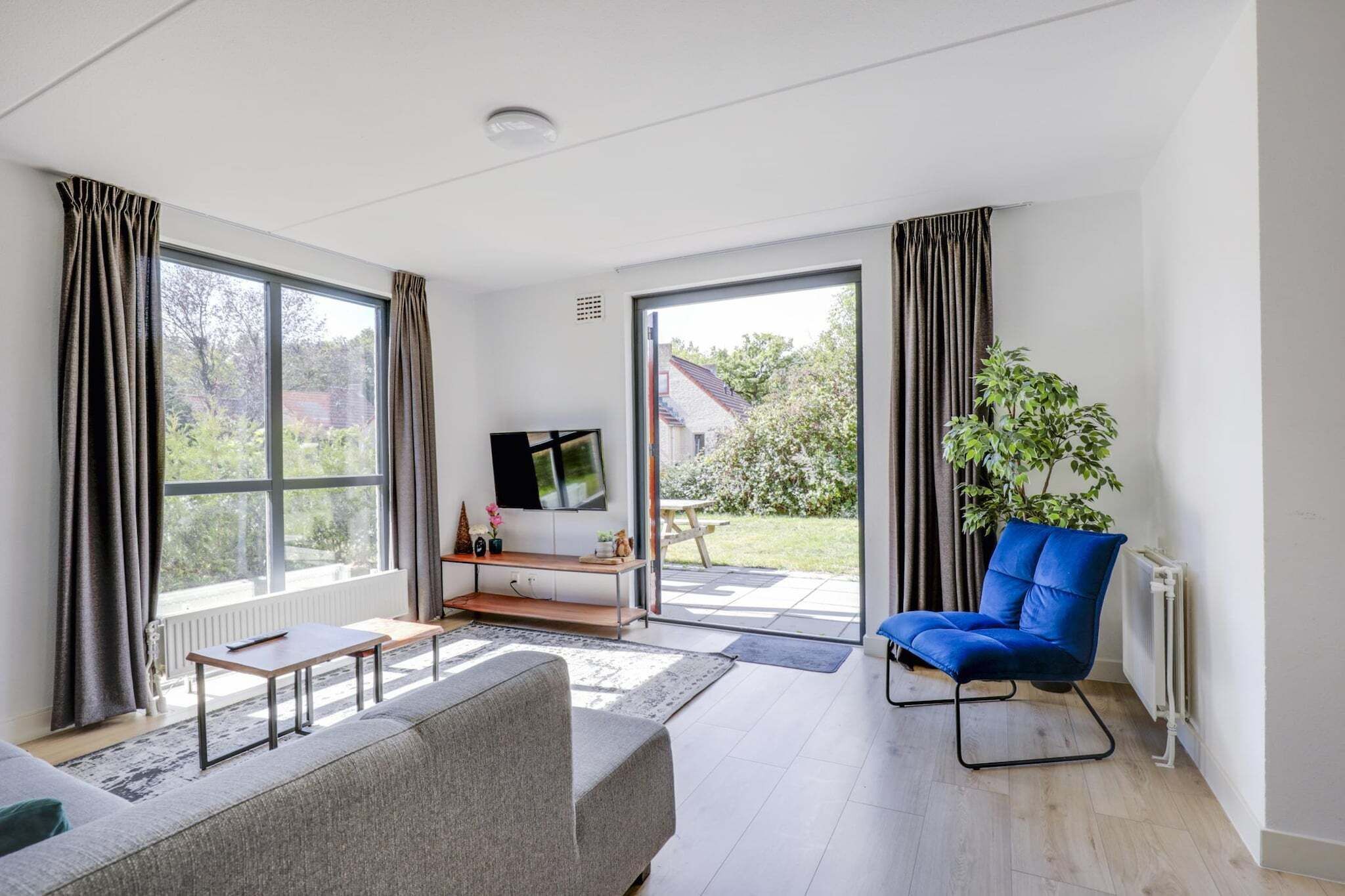 A modern living room with a sofa, a blue chair, a TV on a stand, and a view of the outdoors through large windows.