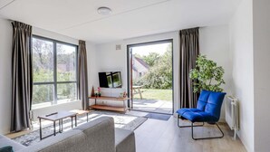 Un salon moderne avec un canapé, un fauteuil bleu, un téléviseur posé sur un meuble et une vue sur l’extérieur grâce à de grandes fenêtres.