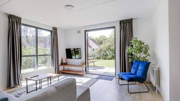 A modern living room with a sofa, a blue chair, a TV on a stand, and a view of the outdoors through large windows.