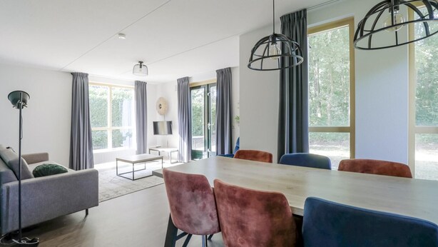 Una sala de estar moderna con sofá, una mesa de centro, una mesa de comedor con sillas y ventanas grandes con cortinas.