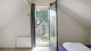 Una habitación con techo inclinado, una cama, un radiador, una ventana con cortinas y vistas a un edificio.
