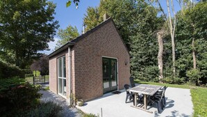 Ein Backsteinhaus mit Terrasse und Tisch, umgeben von Bäumen und Grünpflanzen.