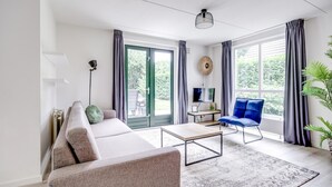 Ein modernes Wohnzimmer mit einer Couch, einem Couchtisch, einem blauen Sessel, einem Fernseher und einem großen Fenster mit Vorhängen.