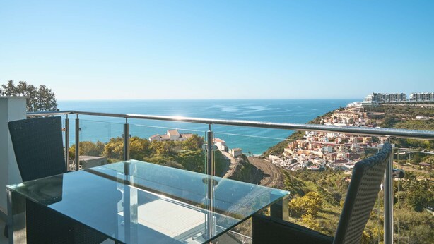 Un balcon avec une table et des chaises en verre, donnant sur une ville côtière et la mer.