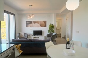 Ein modernes Wohnzimmer mit einer Couch, Sesseln, einem Esstisch, einem Fernseher und einer Pflanze.