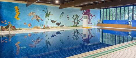 An indoor swimming pool with a mural of marine life on the walls, a diving board, and a bench area.