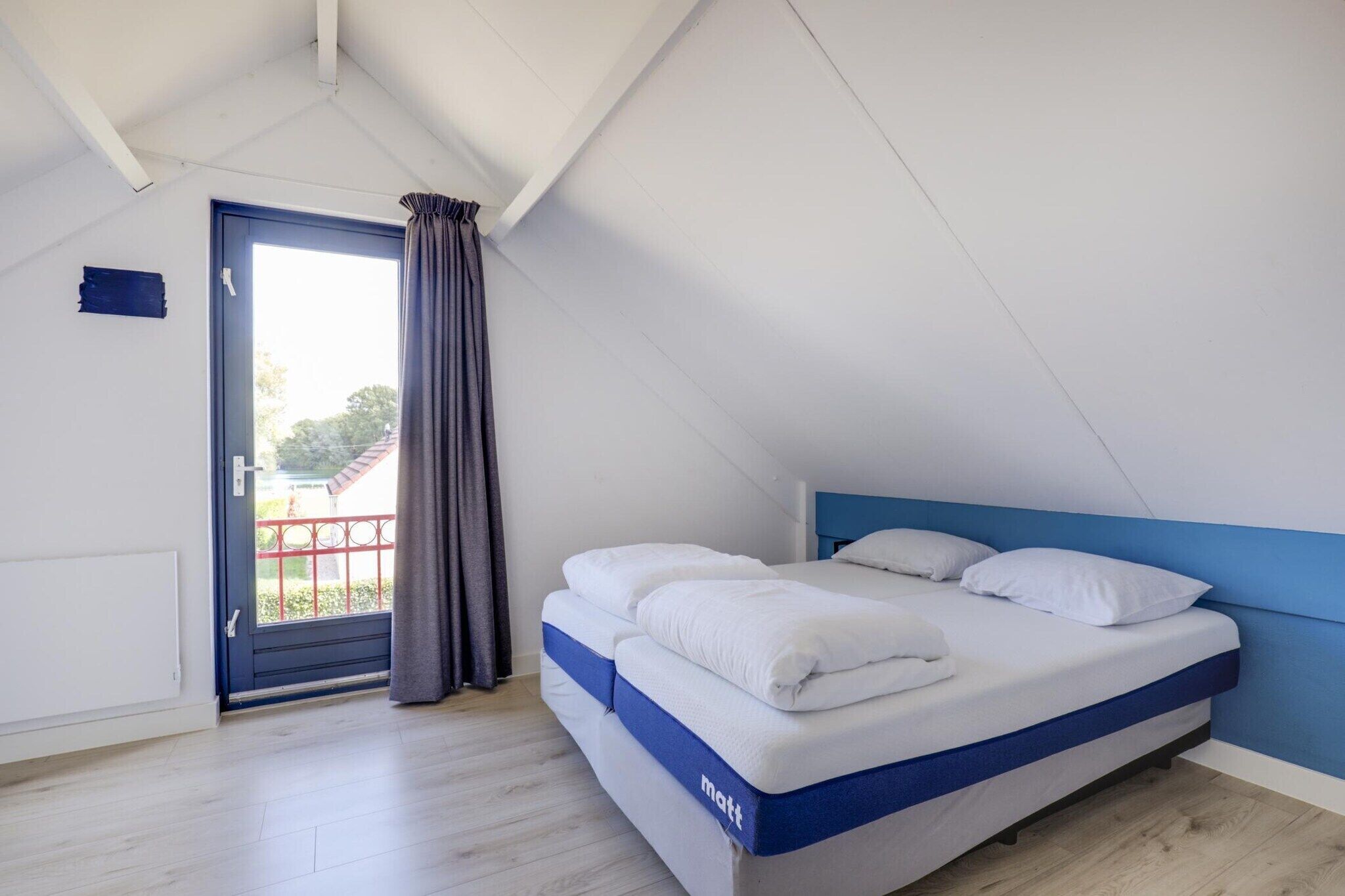 Un dormitorio con techo inclinado, una cama con ropa blanca, un cabecero azul y una puerta que da a un balcón.