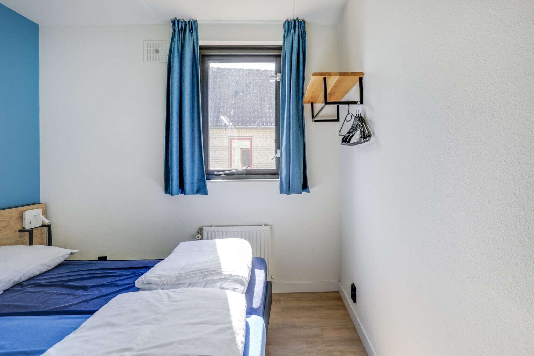 A single bed with a blue bedspread, a white radiator, a window with blue curtains, and a wall-mounted shelf with hanging items.