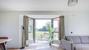 Een moderne woonkamer met een bank, een eettafel en uitzicht op een tuin door grote ramen.