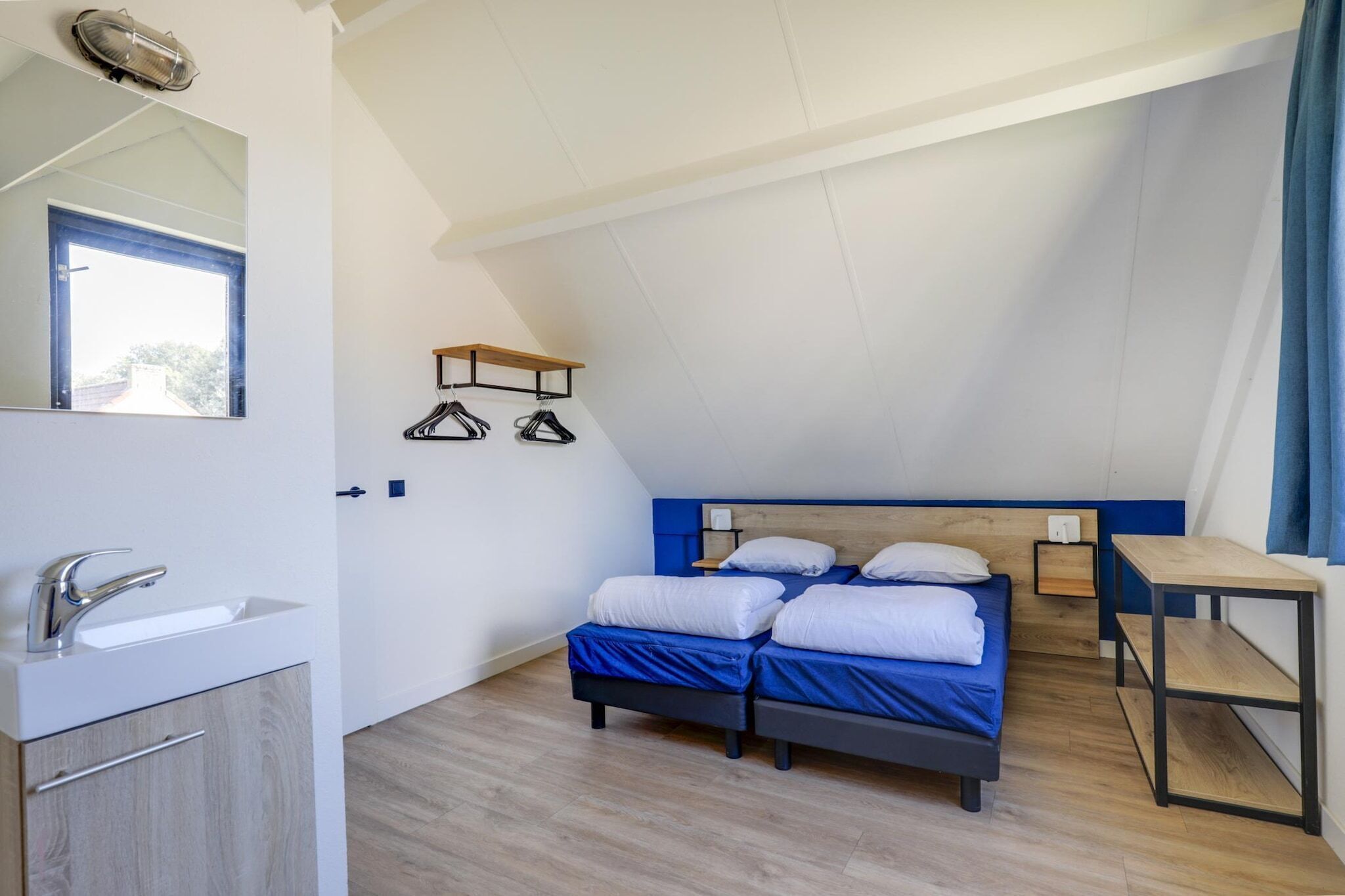 Una camera da letto con soffitto spiovente, un letto con biancheria azzurra, un comodino in legno, uno specchio e un appendiabiti.