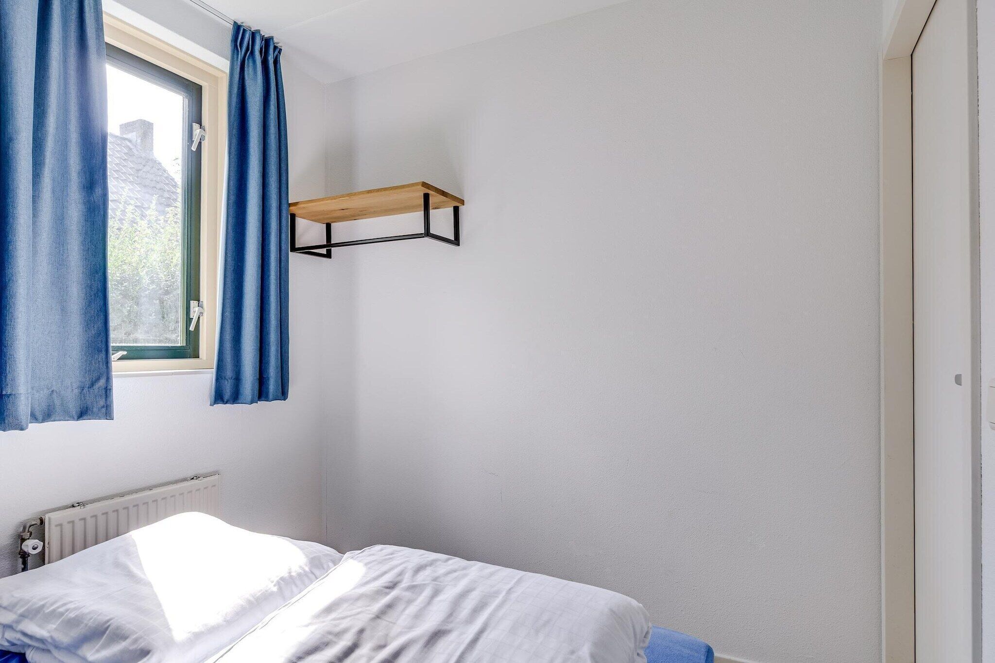 Un dormitorio con cama, ventana con cortinas, radiador y estante de madera.