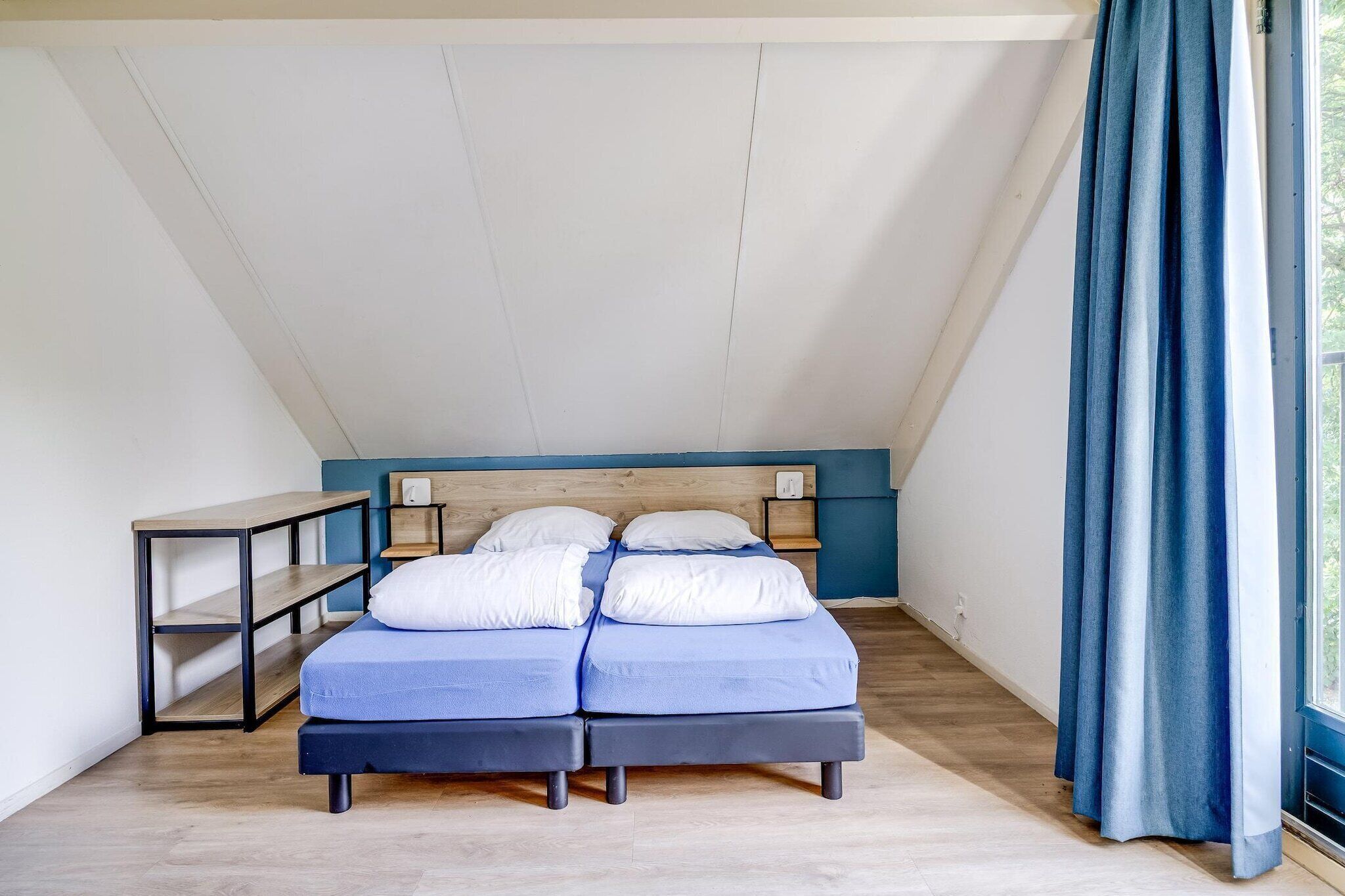 A bedroom with a sloped ceiling, a bed with blue bedding, a bedside table, and a window with curtains.