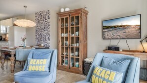 Ein Wohnzimmer mit zwei blauen Sesseln, einer Holzkabine, einem Essbereich und einem Fernseher, der eine Strandansicht zeigt.