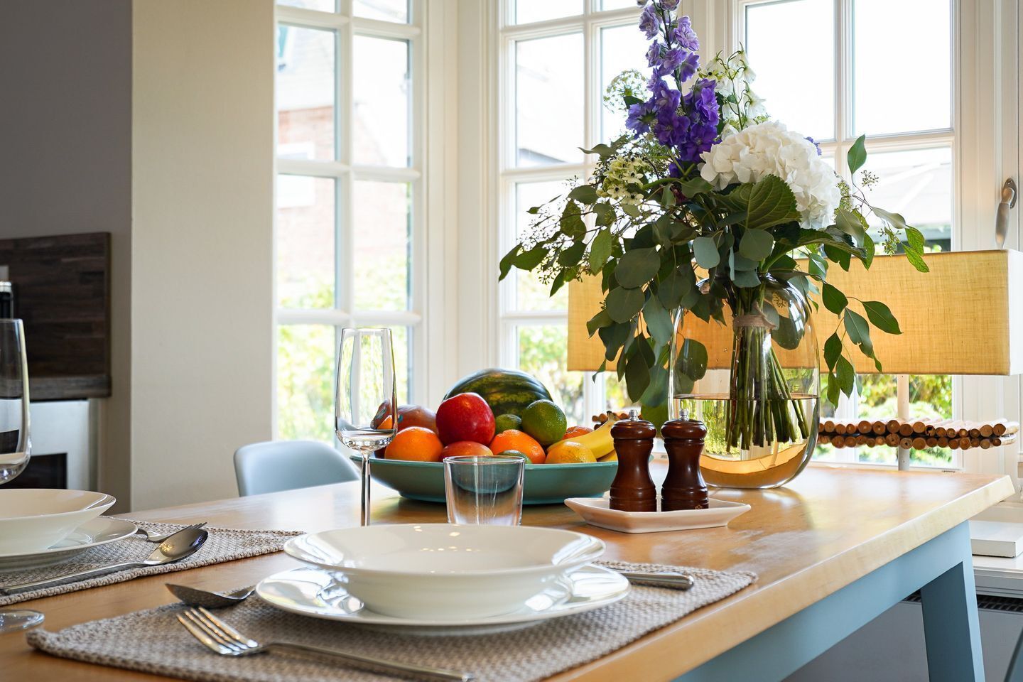 Et spisebord med tallerkener, bestik, en skål med frugt, en vase med blomster og et vindue med udsigt.